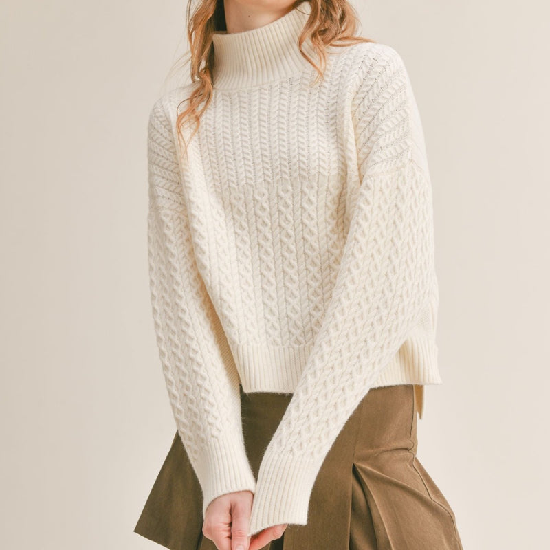Front view of model wearing sweater. Shows the turtle neck neckline and the cable knit detail. Also shows the front is shorter. 