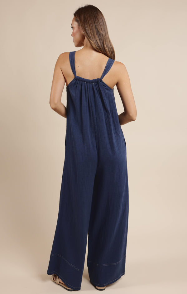 back view of the model wearing the breezy lane jumpsuit. shows the frayed hem. also shows the straps, the wide leg and the white stitching.