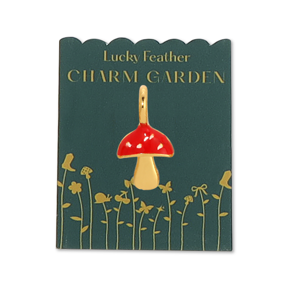Front view of the gold mushroom charm on green packaging with gold wording LUCKY FEATHER CHARM GARDEN and gold flowers on the bottom. The gold mushroom charm has a red top with little speckles of gold on it. 