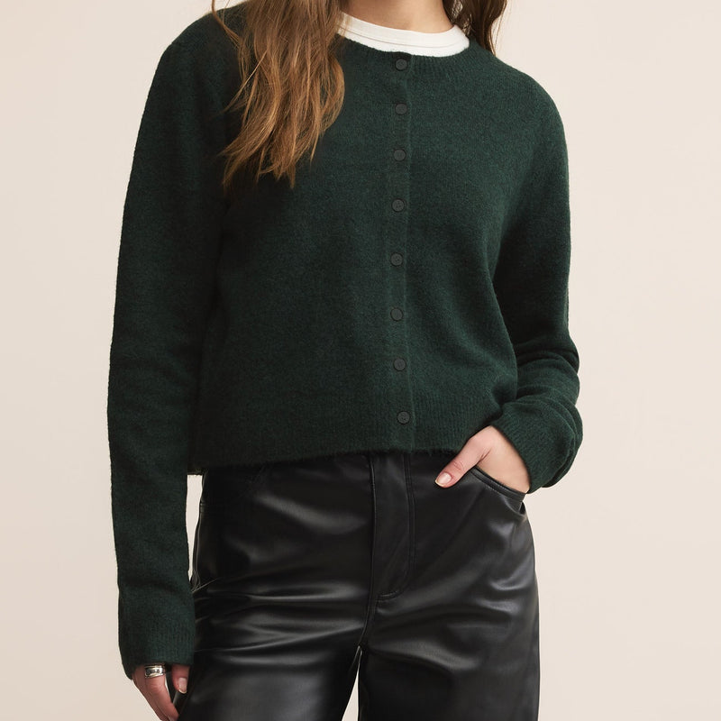 Woman wearing a dark green cardigan over a white shirt and black pants on a beige background