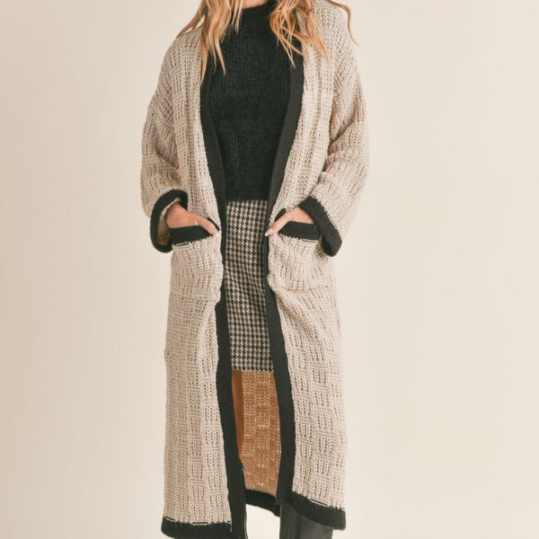 front view of model wearing the Zenni cardigan in taupe. shows the black trim detail. also shows the two front oversized pocket, the oversized sleeves, and the knit sweater detailing. 