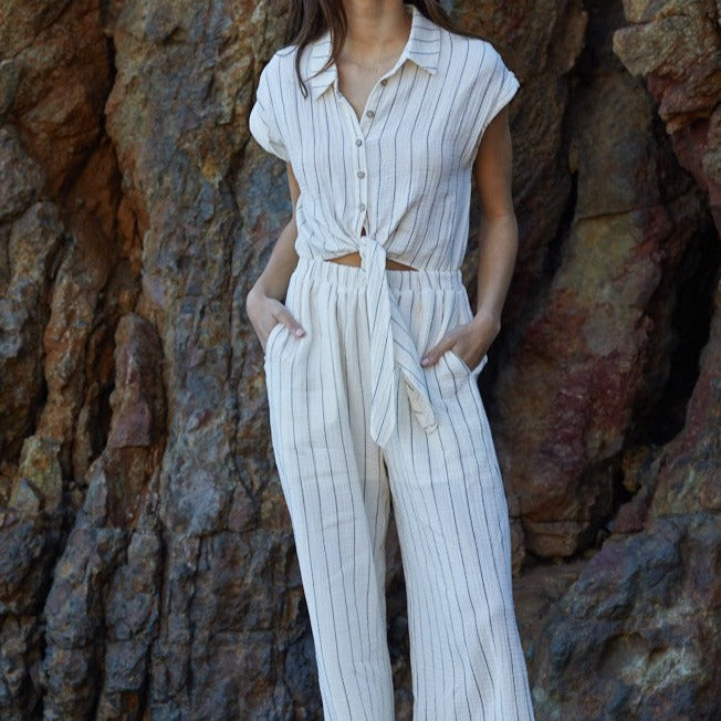 shows  front view of model wearing the jumpsuit. features a collared neckline, buttons down the front that lead into a tie detail. has a cutout feature under the tie and an elastic waistband. has side pockets.