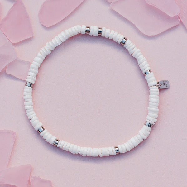 shows a pink backdrop under a stretch bracelet with white and silver flat beads. features a charm etched with "pura vida"