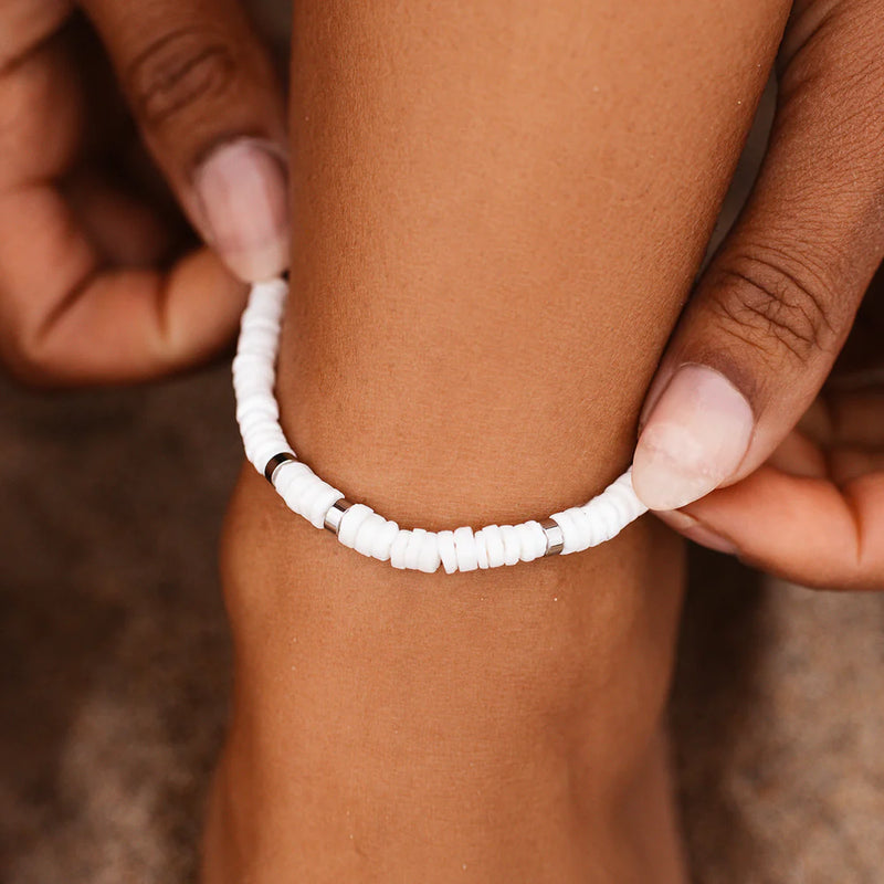 shoiws model holding the stretch anklet on ankle. anklet features white and silver flat beads.